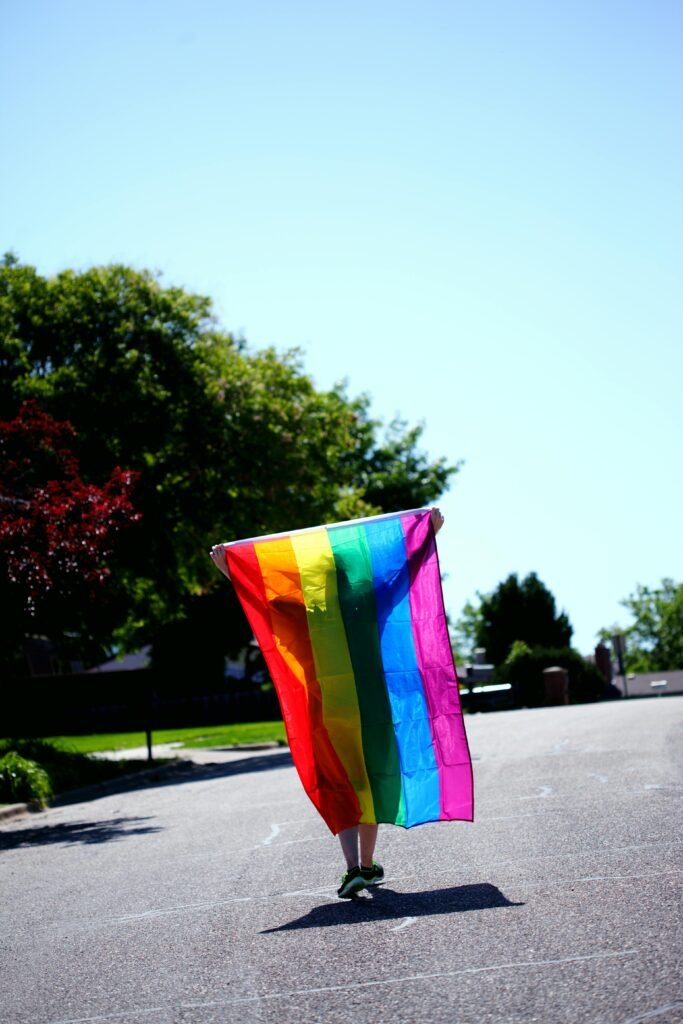 A vibrant pride flag held aloft against a sunny suburban street background.